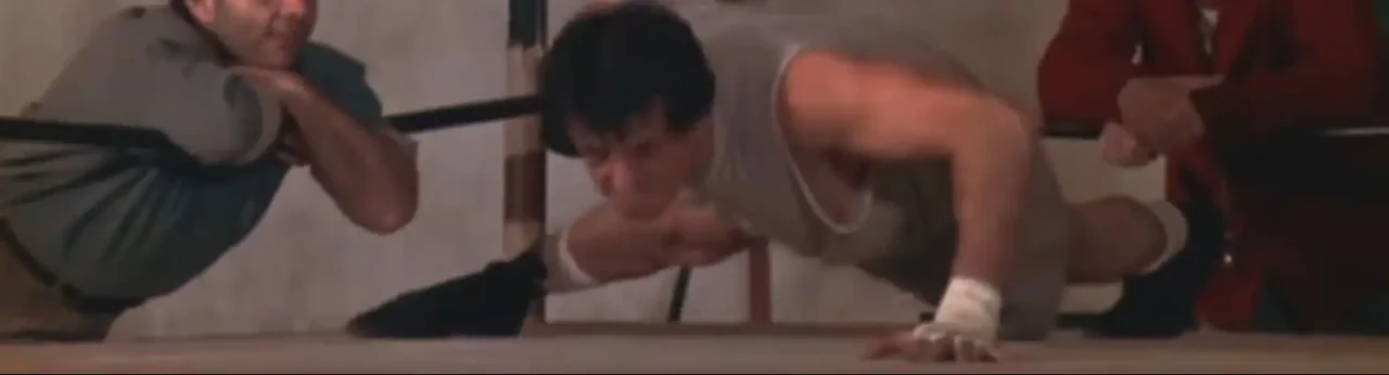 Rocky doing pushups with coach and team mate looking outside the boxing ring
